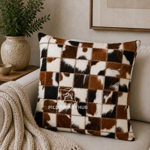 May include: A square throw pillow featuring a patchwork design of cowhide squares in shades of brown, black, and white. The center of the pillow displays a white logo with the text THE PILLOW ART HUB and a small graphic of a paintbrush.