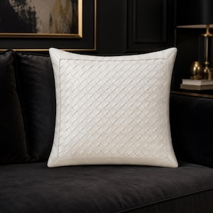 May include: A square throw pillow featuring a cream-colored exterior with a woven, basket-weave pattern across the front surface. The pillow has a smooth, solid border frame that outlines the central woven design, creating a structured appearance.