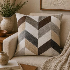 May include: Square decorative pillow with a chevron pattern in shades of brown, beige, and gray. The pillow is on a beige armchair, with a neutral-toned background. The pillow has a soft, textured appearance.