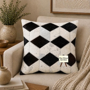 Op de afbeelding: Vierkant decoratief kussen met een geometrisch ruitpatroon in zwart en wit. Het kussen is gemaakt van een textuurrijk materiaal en ligt op een beige fauteuil. Het logo The Pillow Art Hub is zichtbaar.