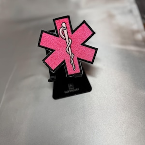 Peut inclure: Écusson rose brodé en forme d'étoile de vie avec une bordure noire et un symbole de caducée rose clair. L'écusson est présenté sur un support de téléphone noir sur fond argenté. L'écusson est un symbole des services médicaux d'urgence.