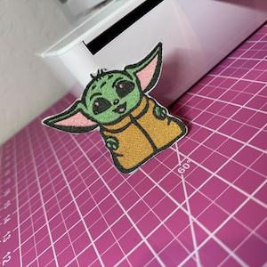 May include: An embroidered patch featuring a green creature with large dark eyes, pointed ears with pink inner details, and a tan robe. The patch has a black stitched border and is positioned against a white object and a pink cutting mat.