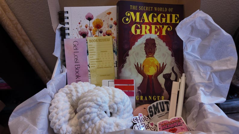 May include: A gift box containing a book, notebook, and other items. The book is titled "The Secret World of Maggie Grey". There is also a white crocheted item, stickers, and a bookmark that says "Get Losi Book".