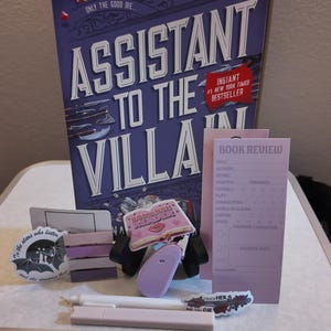 Puede incluir: Un conjunto de art&iacute;culos tem&aacute;ticos de libros, que incluyen un libro de tapa dura titulado "Assistant to the Villain", pegatinas, una hoja de revisi&oacute;n de libros y un bol&iacute;grafo. El libro es morado y blanco, y las pegatinas y la hoja son de tonos morados y rosas.
