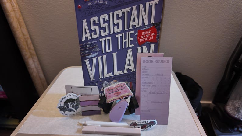 Puede incluir: Un conjunto de art&iacute;culos tem&aacute;ticos de libros, que incluyen un libro de tapa dura titulado "Assistant to the Villain", pegatinas, una hoja de revisi&oacute;n de libros y un bol&iacute;grafo. El libro es morado y blanco, y las pegatinas y la hoja son de tonos morados y rosas.