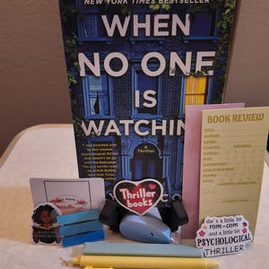 May include: A book titled "When No One Is Watching" is the focal point, accompanied by a "Thriller books" heart-shaped sticker, a book review card, and other small items. The book cover features a blue building illustration.