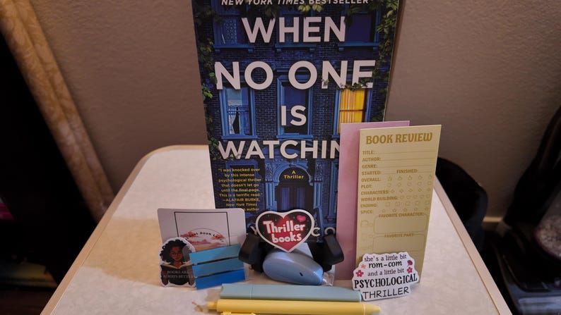 Puede incluir: Un libro titulado "When No One Is Watching" es el punto focal, acompa&ntilde;ado de una pegatina en forma de coraz&oacute;n "Thriller books", una tarjeta de rese&ntilde;a de libro y otros objetos peque&ntilde;os. La portada del libro presenta una ilustraci&oacute;n de un edificio azul.