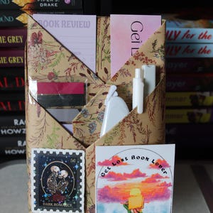 Puede incluir: Un organizador de libros hecho a mano con papel estampado floral. Cuenta con bolsillos que contienen un bloc de notas de rese&ntilde;as de libros, pegatinas y una peque&ntilde;a botella blanca. Una pegatina dice "Get Lost Book Lover."