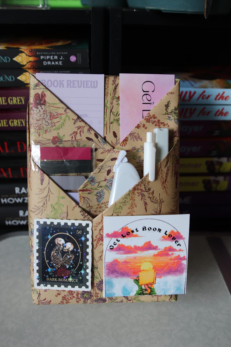 Puede incluir: Un organizador de libros hecho a mano con papel estampado floral. Cuenta con bolsillos que contienen un bloc de notas de rese&ntilde;as de libros, pegatinas y una peque&ntilde;a botella blanca. Una pegatina dice "Get Lost Book Lover."