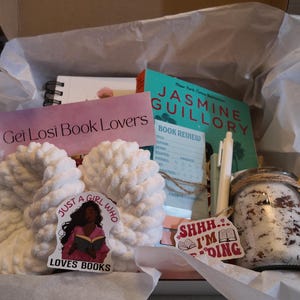 May include: A gift box filled with a variety of items for book lovers. Includes a book, a notebook, a jar, a pen, a pair of white slippers, and stickers. The box is lined with white tissue paper. The text on the sticker says "Just a girl who loves books."