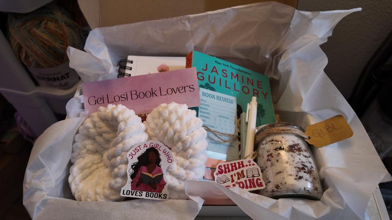 May include: A gift box filled with a variety of items for book lovers. Includes a book, a notebook, a jar, a pen, a pair of white slippers, and stickers. The box is lined with white tissue paper. The text on the sticker says "Just a girl who loves books."