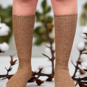 May include: A pair of knee-high socks in a warm, tan color. The socks have a ribbed knit texture and are shown from the knees down, with a blurred background of cotton branches.