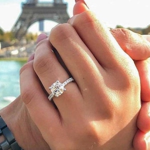 Peut inclure: Une bague de fiançailles en diamant avec une grosse pierre centrale de taille ronde et un anneau pavé. La bague est portée au doigt, tenue par la main d'une autre personne. La Tour Eiffel est visible en arrière-plan.