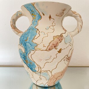 May include: A white ceramic vase with two handles, decorated with blue, brown, and gold abstract map-like designs. The vase has a wide mouth and a rounded body. The gold accents highlight the shapes and patterns on the vase.