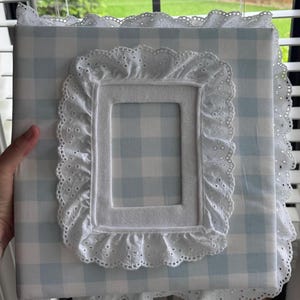Può includere: Un album fotografico a quadretti vichy azzurro e bianco, con cornice e bordi in pizzo sangallo bianco. L'album è rettangolare. La cornice è centrata sulla copertina. Le misure sono in centimetri.