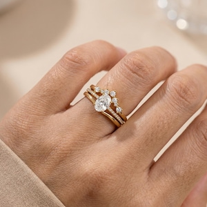 Puede incluir: Un conjunto de tres anillos de oro con detalles de diamantes. Los anillos están apilados en un dedo, mostrando un diamante central de talla ovalada y diamantes más pequeños. Los anillos tienen un diseño delicado y elegante.