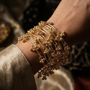 Peut inclure: Une collection de bracelets dorés, chacun orné de petites décorations sphériques. Les bracelets sont empilés sur un poignet, mettant en valeur leur design complexe et leur surface réfléchissante. Les bijoux sont posés sur un fond doux et neutre.