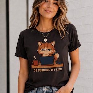 May include: A black short-sleeved t-shirt featuring a graphic of a fluffy orange cat wearing round glasses, sitting at a desk with a laptop and a red coffee mug. The text below the cat reads "DEBUGGING MY LIFE" in a bold, tan-colored font.