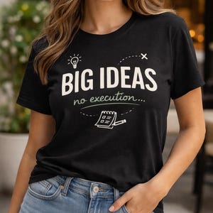 May include: Black t-shirt with the text "BIG IDEAS no execution..." in white. The shirt features a lightbulb graphic and a notepad illustration. The model is wearing blue jeans.