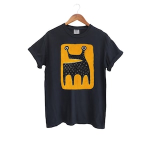 May include: Dark navy blue t-shirt with a unique graphic. The design features a black creature with a textured body, set against a bright yellow rectangular background. The creature has two large eyes and three legs. The t-shirt is hanging on a wooden hanger.