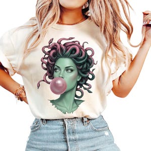 May include: Cream-colored t-shirt featuring a graphic of Medusa blowing a bubblegum bubble. Medusa's face is green with pink and green snakes for hair. The bubble is a light pink color. The shirt has short sleeves.