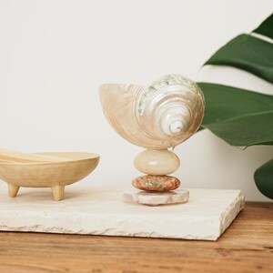 May include: A decorative object featuring a large, iridescent spiral seashell mounted atop a stack of three smooth, rounded stones in shades of cream, mottled red, and translucent white, positioned next to a small, three-legged tan ceramic bowl.