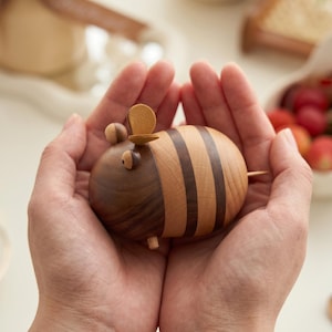 Puede incluir: Una figura de abeja de madera hecha a mano, con un cuerpo a rayas y un pequeño sombrero. La abeja está hecha de madera clara y oscura, con una pequeña cola de madera. La figura se sostiene en manos abiertas.