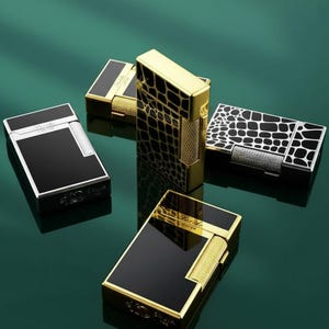 May include: A collection of luxury lighters in different styles. The lighters have black, gold, and silver finishes. Some have a glossy black surface, while others feature textured patterns. The lighters are displayed on a reflective green surface.