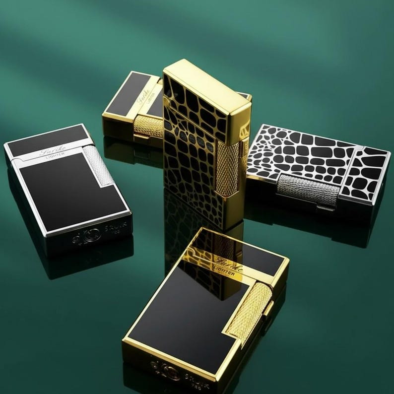 May include: A collection of luxury lighters in different styles. The lighters have black, gold, and silver finishes. Some have a glossy black surface, while others feature textured patterns. The lighters are displayed on a reflective green surface.