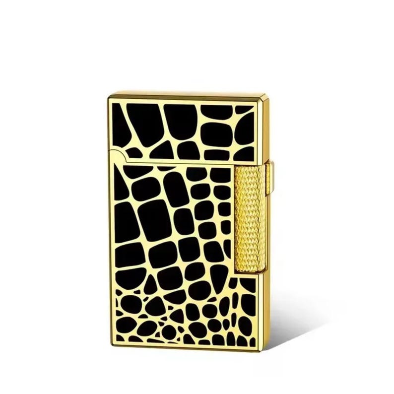 May include: A gold-coloured lighter featuring a black, textured pattern. The lighter has a rectangular shape with a textured side for grip and a hinged top. The design resembles reptile skin.