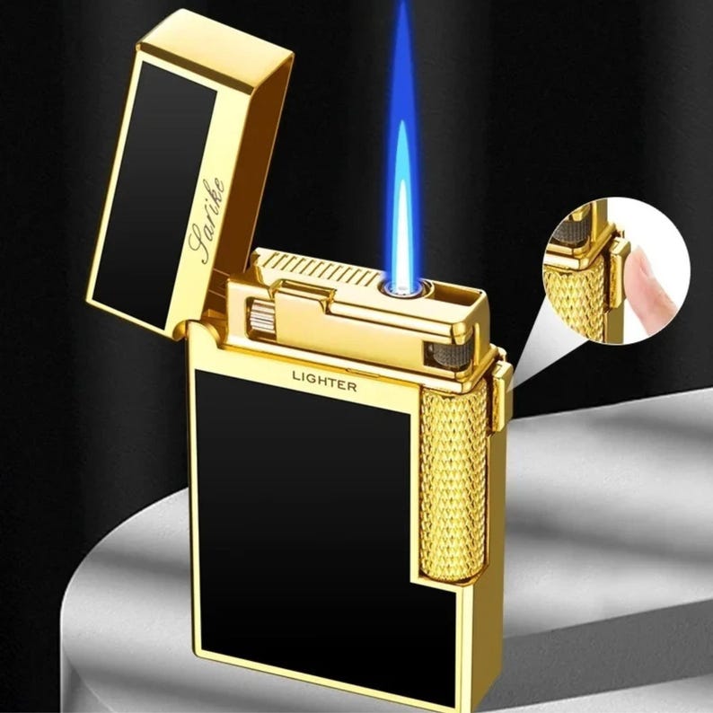 May include: Gold-coloured lighter with a black panel and a blue flame. The lighter has the word "Lighter" printed on it. The brand name "Sarike" is printed on the lid. The lighter is open, with the flame lit.