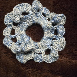 May include: A handmade, blue and white crocheted hair scrunchie. The scrunchie is circular with a central opening and a scalloped edge. The yarn appears to be a blend of light and dark blue, creating a textured appearance. The scrunchie is approximately 4 inches in diameter.