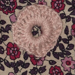 Op de afbeelding: Een lichtroze, gebreide scrunchie met een cirkelvormig ontwerp. De scrunchie is geplaatst tegen een achtergrond van stof met een bloemenpatroon in roze, paarse en beige tinten. De scrunchie is een haaraccessoire.