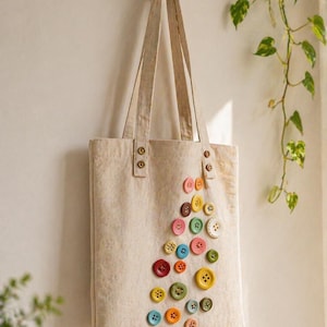 Puede incluir: Un bolso tote beige con una colección de botones coloridos dispuestos en un patrón decorativo. El bolso cuelga de un gancho de madera en una pared blanca, con una planta verde en el fondo.
