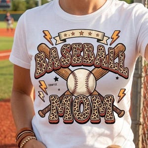 Op de afbeelding: Wit T-shirt met een honkbalontwerp. Het ontwerp bevat de woorden "BASEBALL MOM" in een luipaardprint lettertype, gekruiste honkbalknuppels, een honkbal, sterren en bliksemschichten.