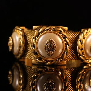 May include: A gold-tone bracelet featuring a wide mesh band adorned with three circular ornaments. Each ornament has a smooth, cream-colored center, a twisted rope-style border, and a small, decorative metallic emblem in the middle.