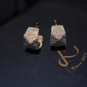 May include: A pair of silver-toned hoop earrings featuring a textured, glittery surface. The earrings have a wide, curved design and are secured with post-back closures. The surface of each hoop reflects light, creating a sparkling appearance.