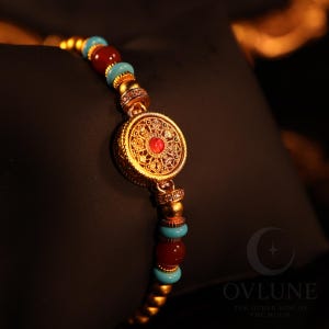 Könnte beinhalten: Ein goldfarbenes Armband mit einem zentralen runden Anhänger, der filigrane Muster und einen kleinen roten Stein in der Mitte aufweist. Das Band besteht aus abwechselnd glatten roten Perlen, hellen türkisfarbenen Perlen und kleinen goldenen Zwischenelementen.