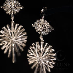 May include: A pair of silver-toned drop earrings featuring a textured, starburst-like design. Each earring consists of a smaller, multi-faceted geometric top piece connected to a larger, circular, radiating sunburst pendant with thin, metallic spokes.