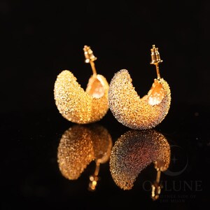 May include: A pair of gold-toned hoop earrings with a textured, granular surface. The earrings are semi-circular in shape and feature a post-back closure mechanism. The design has a reflective, sparkling appearance against a dark background.