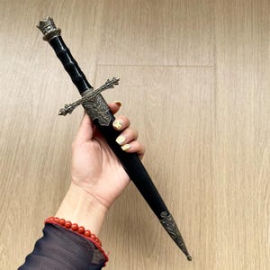 Puede incluir: Una daga decorativa con mango negro y una vaina negra a juego. La empuñadura y la contera de la vaina están hechas de metal en tono bronce antiguo con intrincados detalles en relieve, incluyendo una figura alada en la cruz.