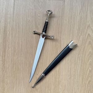 Pode incluir: Uma adaga de metal de cor prateada com lâmina pontiaguda, guarda transversal e cabo preto canelado. A adaga é acompanhada por uma bainha preta a condizer com detalhes em metal prateado na ponta e na abertura da bainha.