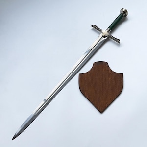 Peut inclure: Une longue épée en acier à double tranchant avec une pointe acérée, une garde en argent ornée de détails métalliques complexes et un manche cylindrique vert foncé surmonté d'un pommeau en argent, avec une plaque murale en bois.