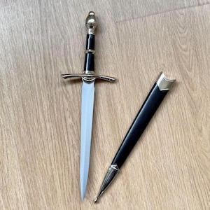 May include: A silver-toned dagger with a pointed blade, a black handle, and a rounded pommel. The crossguard is thin and silver-toned. Beside the dagger lies a matching black scabbard featuring a silver-toned metal tip and a silver-toned throat.