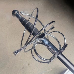 Pode incluir: Uma espada do tipo florete com uma guarda de cesto em cromo polido, apresentando múltiplos anéis circulares para proteger a mão. O punho é revestido em material preto com detalhes em fio prateado, com um pomo redondo e uma guarda reta.