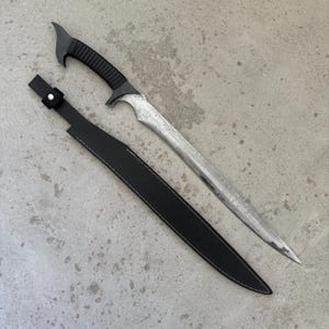 May include: A curved steel blade with a textured, metallic finish and a black ribbed handle. The sword is accompanied by a black sheath featuring a single silver-toned metal stud near the opening, designed to hold the blade securely in place.
