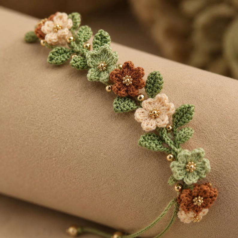 Pu&ograve; includere: Un braccialetto all'uncinetto caratterizzato da una serie di piccoli fiori a cinque petali nelle tonalit&agrave; crema, verde salvia e marrone. Gli elementi floreali sono intervallati da minuscole perline metalliche dorate e foglie all'uncinetto.