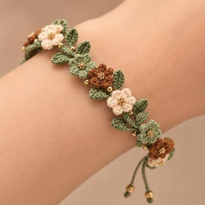 Pu&ograve; includere: Un braccialetto all'uncinetto caratterizzato da una serie di piccoli fiori a cinque petali nei colori crema, verde salvia e marrone. Gli elementi floreali sono intervallati da foglie verdi all'uncinetto e minuscole perline dorate metallizzate.
