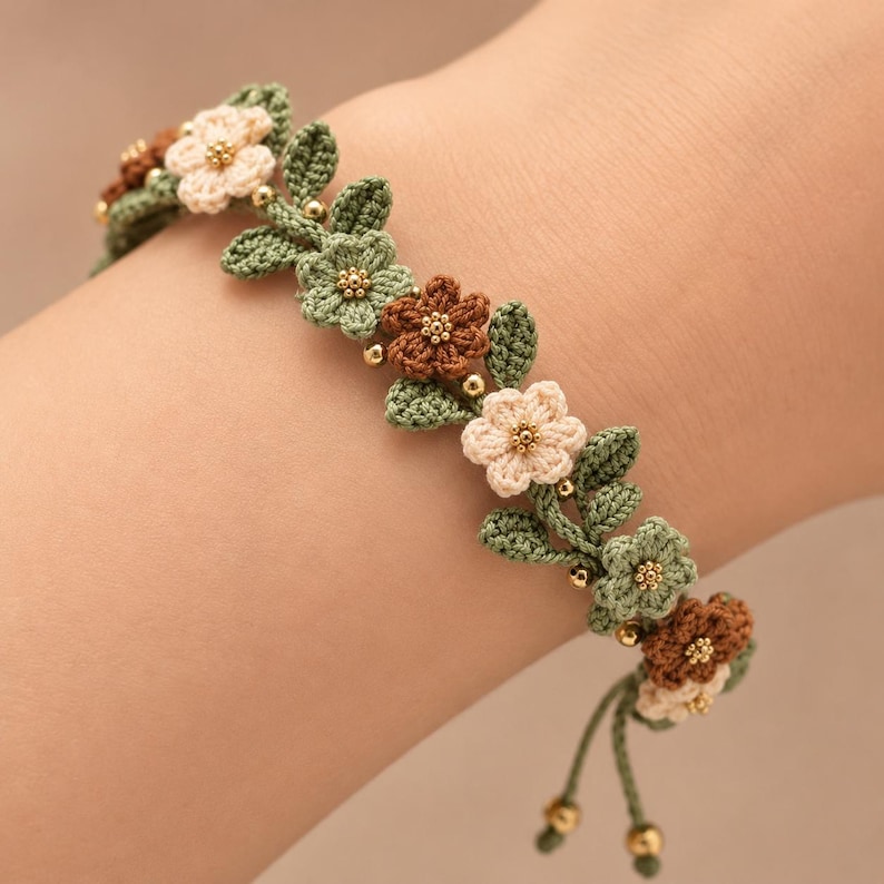 Pu&ograve; includere: Un braccialetto all'uncinetto caratterizzato da una serie di piccoli fiori a cinque petali nei colori crema, verde salvia e marrone. Gli elementi floreali sono intervallati da foglie verdi all'uncinetto e minuscole perline dorate metallizzate.