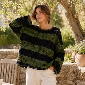 May include: A long-sleeved crochet sweater featuring wide horizontal stripes in alternating dark forest green and black. The garment has a loose, open-knit construction with a round neckline and a relaxed fit, paired with plain white trousers.